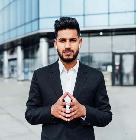 Portrait of a serious smiling modern Indian man, businessman, in front of the holding, behind him a modern building, he is looking at the camera